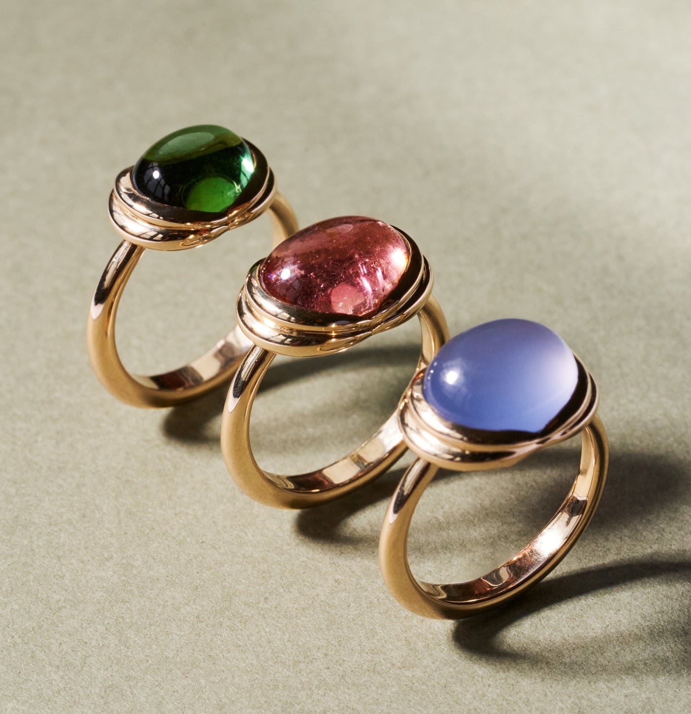 Three gold rings with oval cabochon gemstones in green, pink, and blue, arranged diagonally on a textured sage green surface.