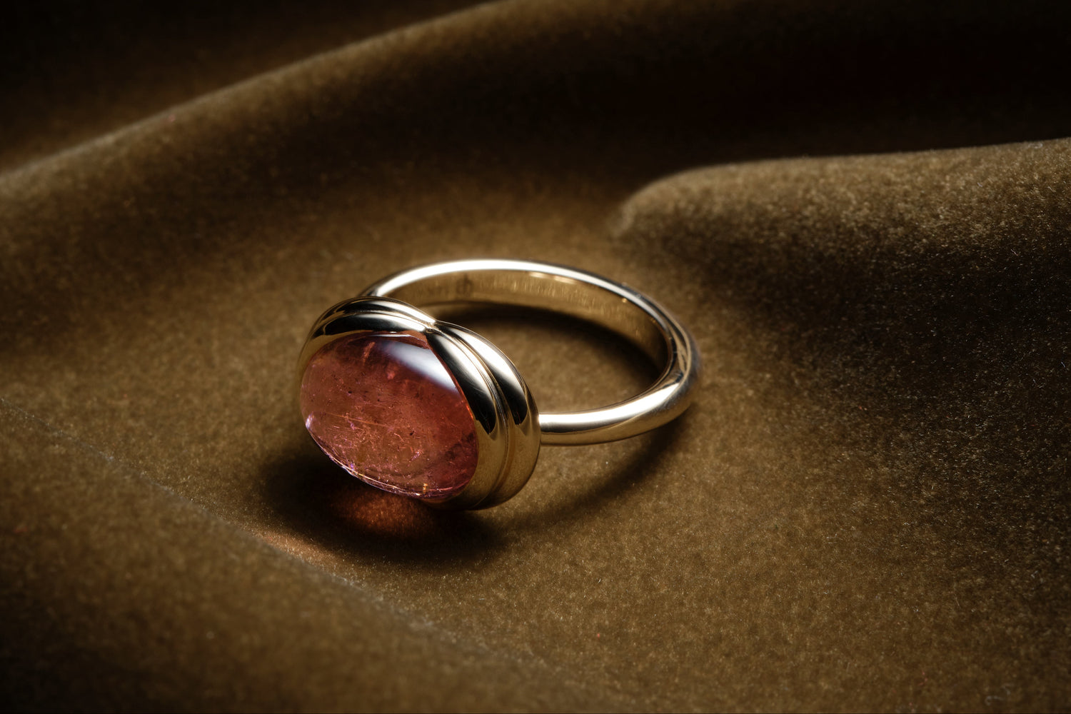 A gold ring with an oval, polished blush colored gemstone, resting on draped dark brown fabric.