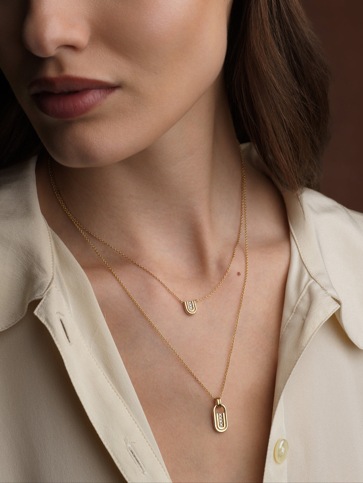 A close-up of a woman wearing layered gold necklaces, including a small oval shaped pendant and a delicate chain necklace, paired with a cream-colored collared shirt.