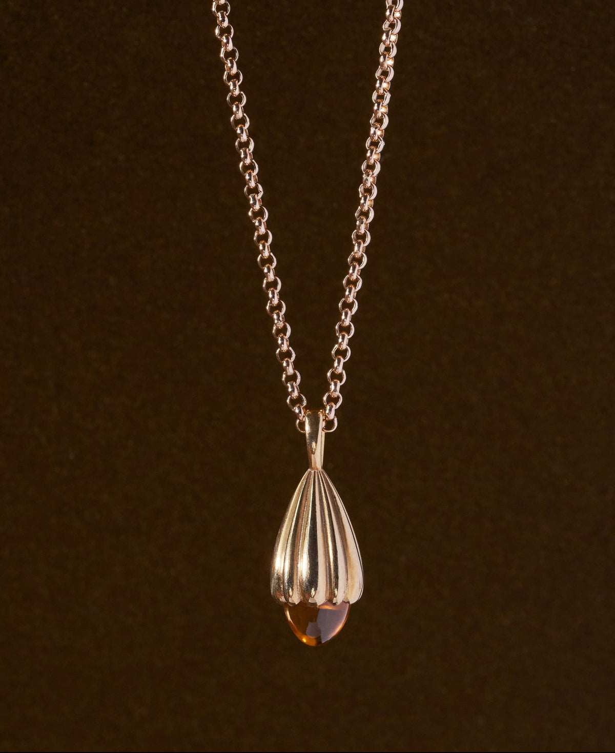 Gold necklace featuring a teardrop-shaped pendant with fluted ridges and a faceted amber-colored stone at the bottom, set against a dark brown background.