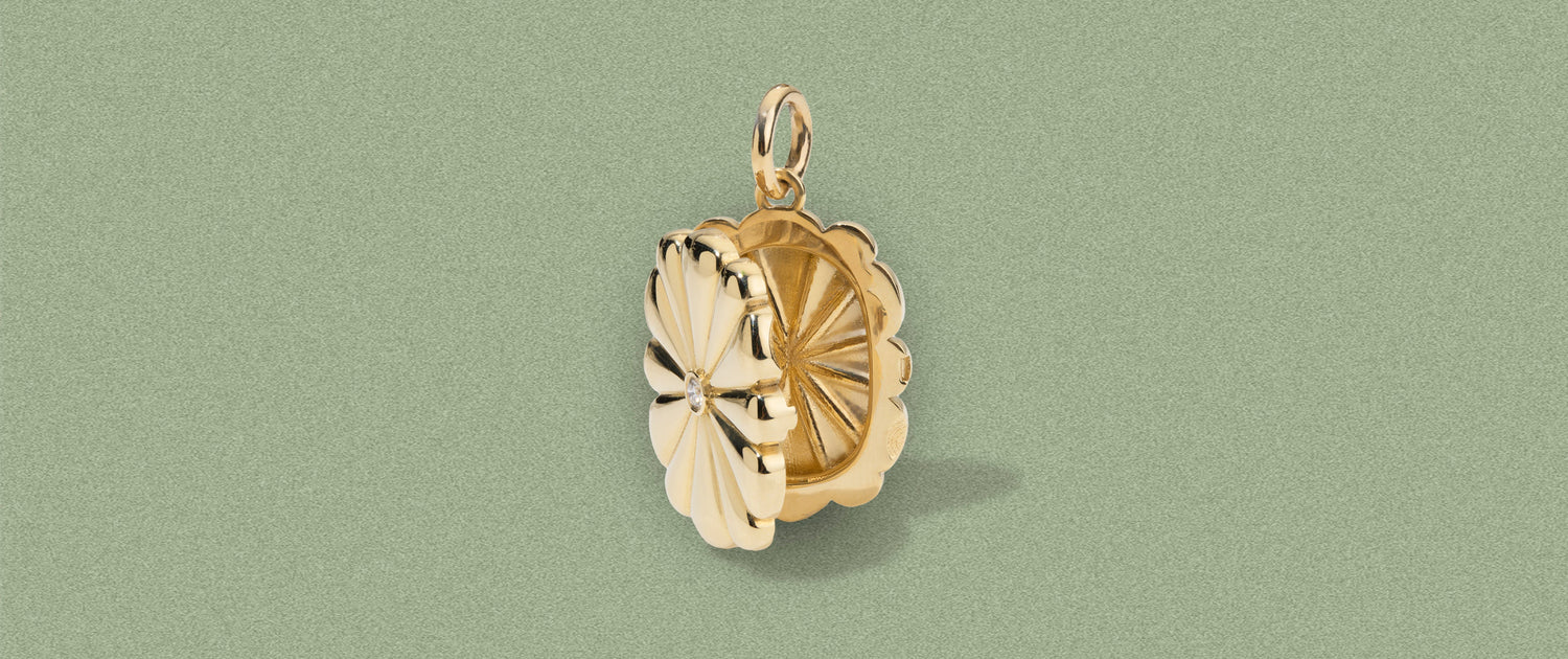 A gold-toned flower-shaped pendant with a textured center and a small hanging loop at the top, shown on a muted sage green background.