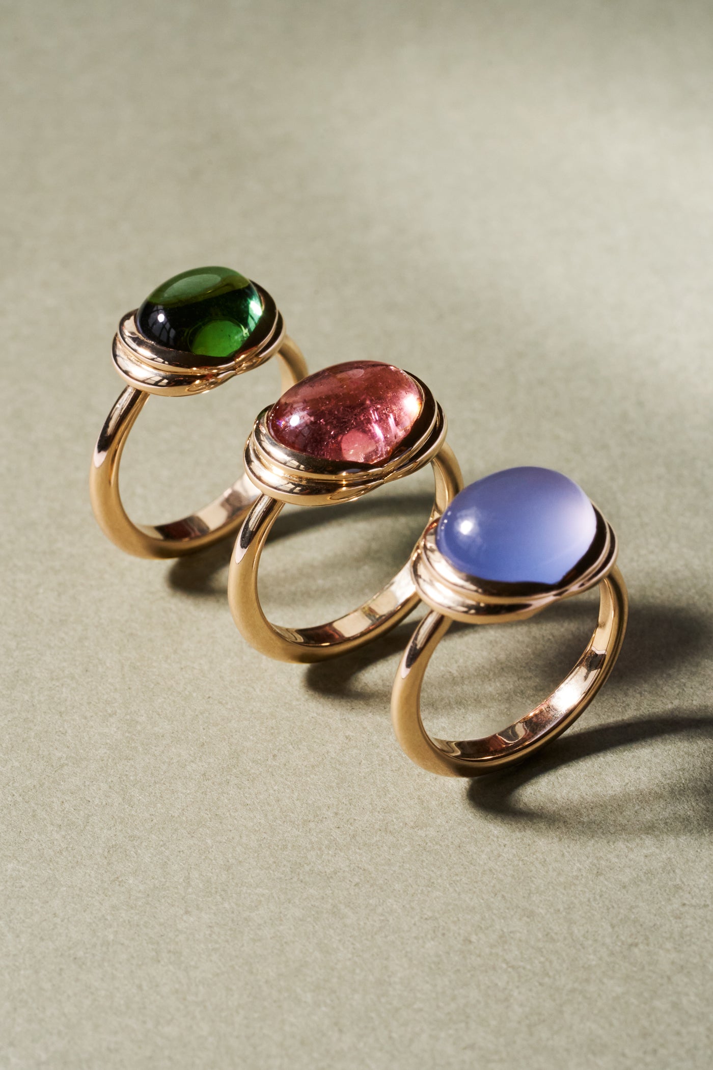 Three gold rings with oval cabochon gemstones in green, pink, and blue, arranged diagonally on a textured sage green surface.