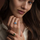 A close-up lifestyle shot of a woman wearing two rings: a silver ring with a light blue oval stone on her middle finger and a silver ring with a green oval stone on her ring finger. She is wearing a cream-colored collared shirt against a dark brown background.