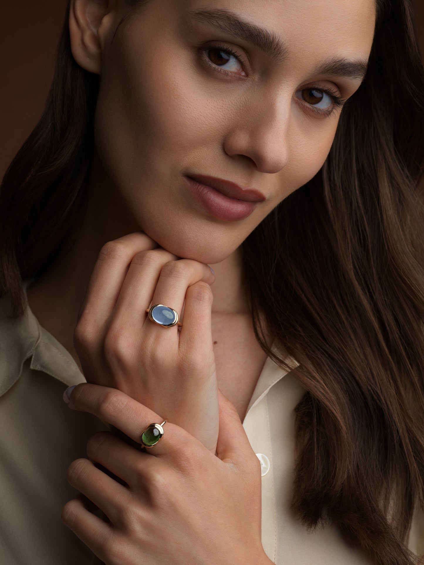 A close-up lifestyle shot of a woman wearing two rings: a silver ring with a light blue oval stone on her middle finger and a silver ring with a green oval stone on her ring finger. She is wearing a cream-colored collared shirt against a dark brown background.