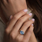 A close-up lifestyle shot of a woman wearing two rings: a gold band ring with a black rectangular stone on one finger, and a gold band ring with a round light blue stone on the other.