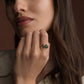 A close-up lifestyle shot of a woman wearing a gold ring with a green oval stone, resting her chin on her hand against a warm brown background.