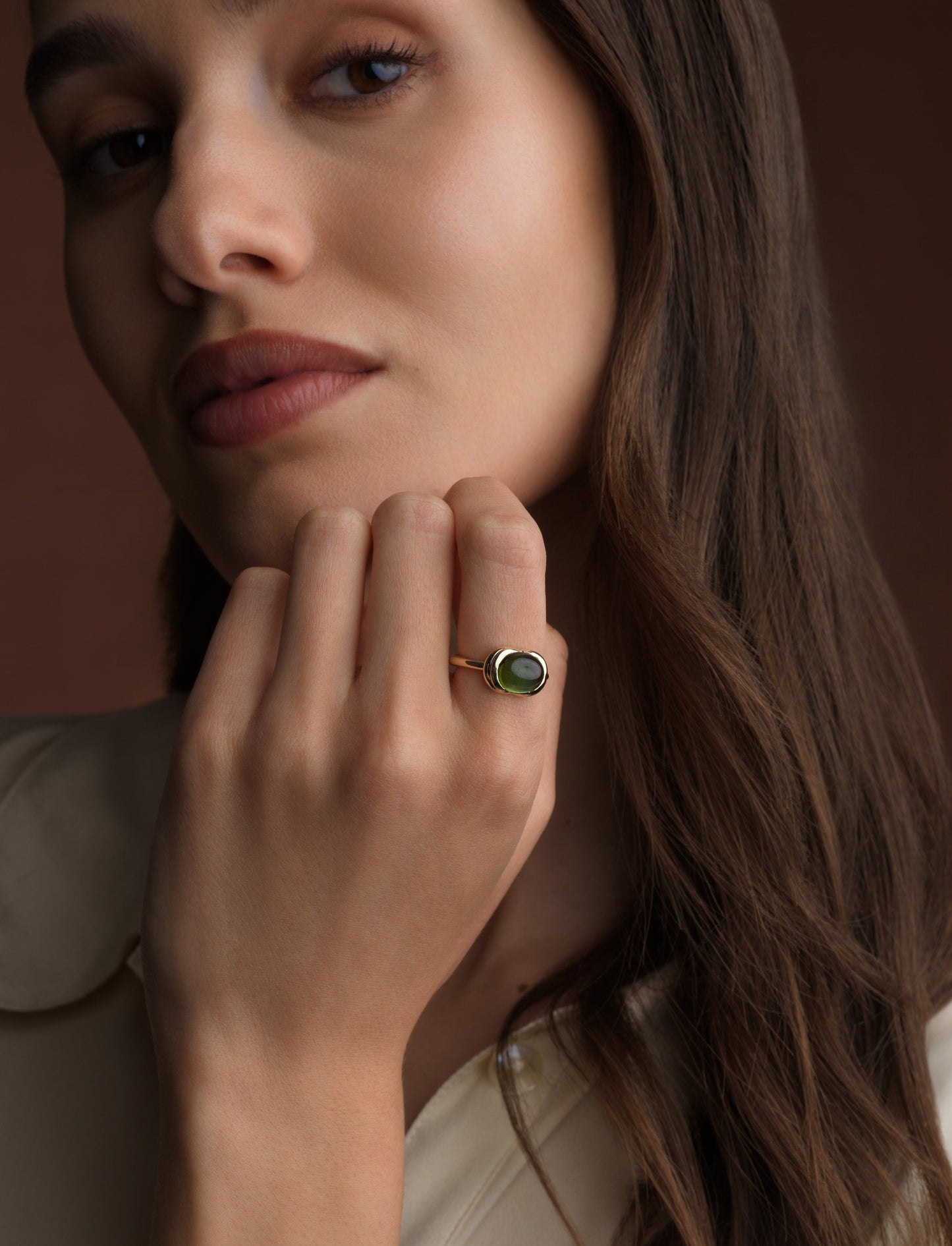 A close-up lifestyle shot of a woman wearing a gold ring with a green oval stone, resting her chin on her hand against a warm brown background.