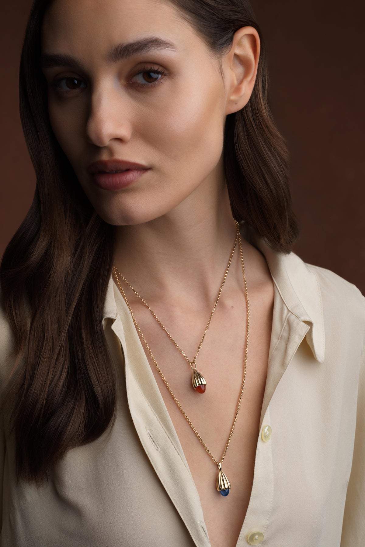 A close-up of a woman wearing layered gold necklaces with teardrop-shaped pendants, one featuring a red stone and the other a blue stone, paired with a cream-colored blouse against a dark brown background.