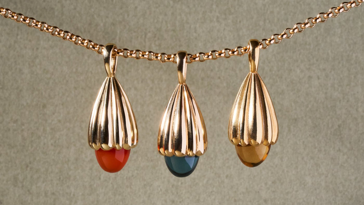 A gold chain necklace featuring three teardrop-shaped pendants with vertical fluting, each holding a small dome-colored stone in red, blue, and gold, displayed against a textured sage green background.