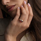 A close-up lifestyle shot of a woman wearing a gold ring with a pink oval  shaped gemstone, resting her hand on her chin against a warm brown background.