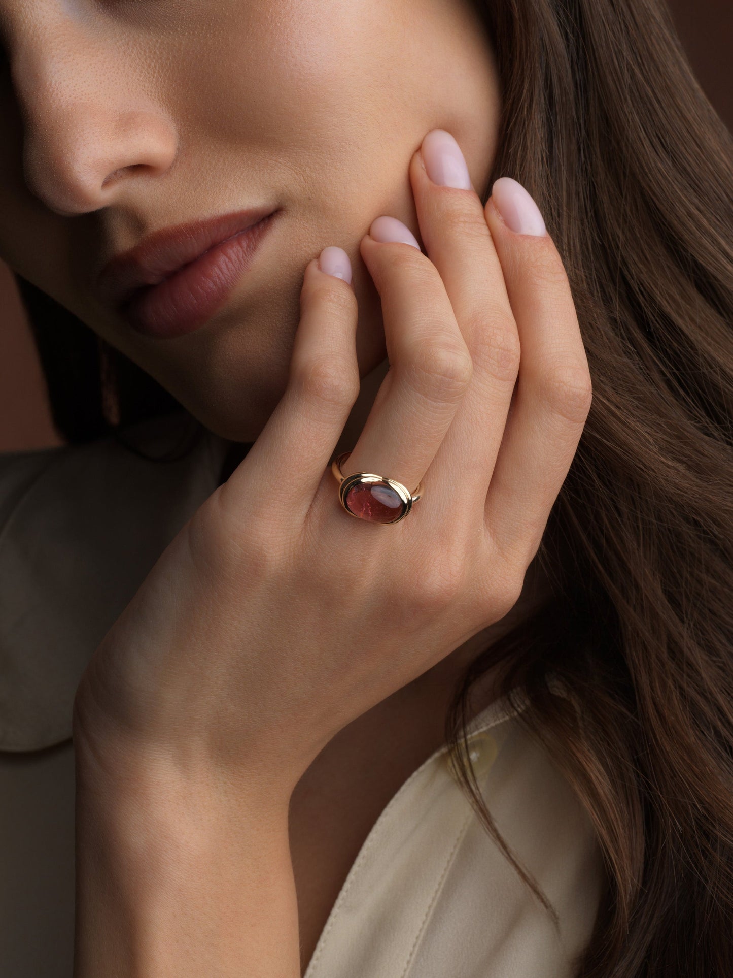 A close-up lifestyle shot of a woman wearing a gold ring with a pink oval  shaped gemstone, resting her hand on her chin against a warm brown background.