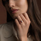 A close-up lifestyle shot of a woman wearing a gold ring with an oval pink gemstone, set against a warm brown background.