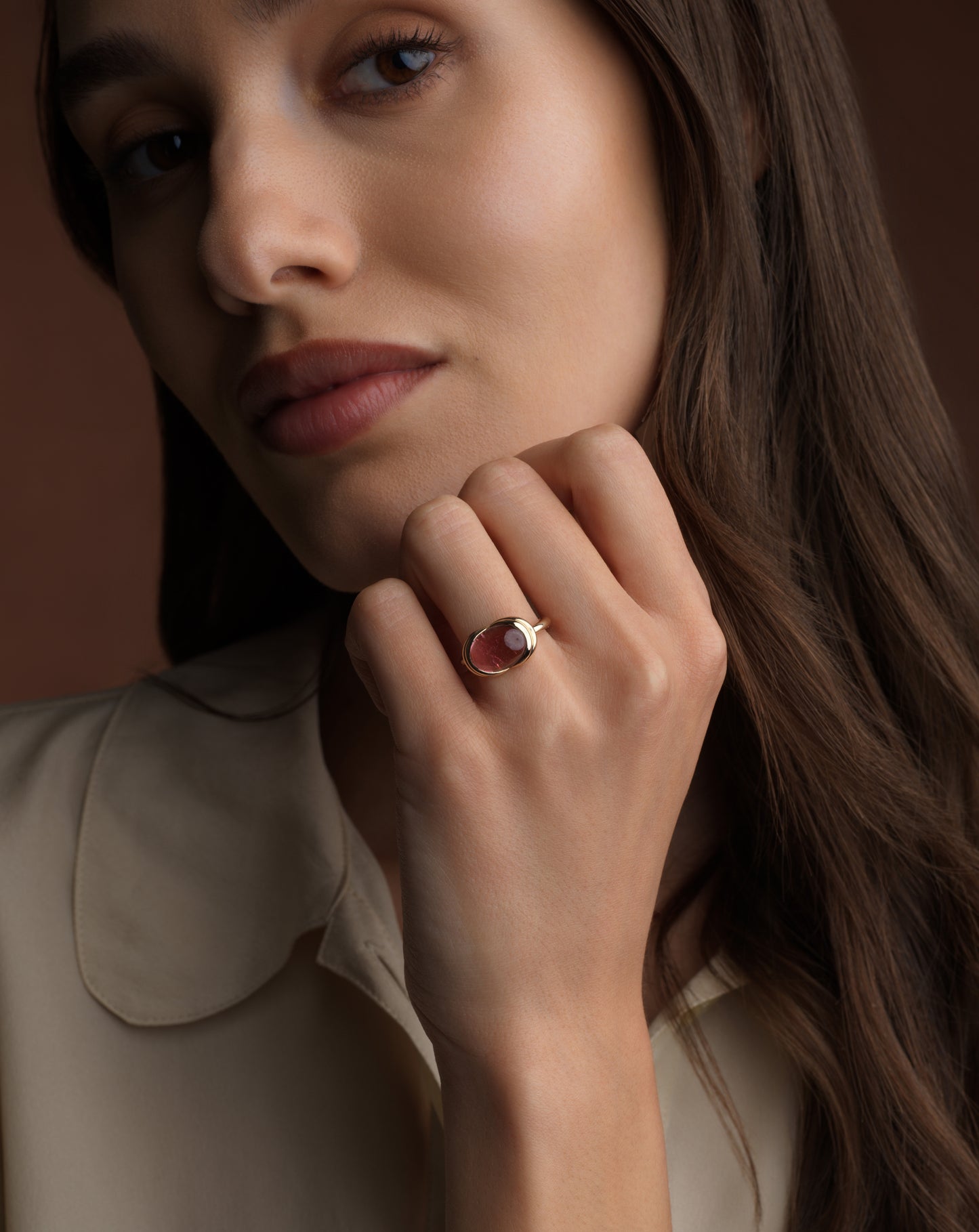 A close-up lifestyle shot of a woman wearing a gold ring with an oval pink gemstone, set against a warm brown background.